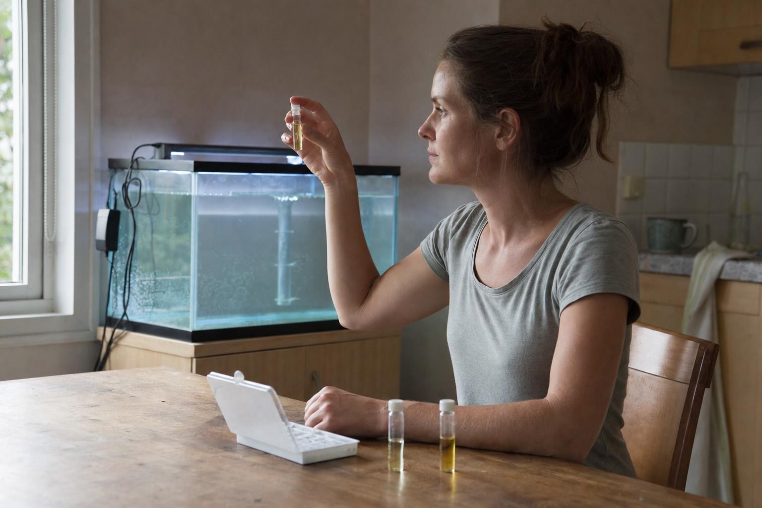 A person at a kitchen table holds a water test tube up to the window light, with an empty cycling aquarium glowing in the background